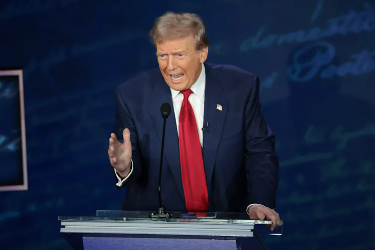 A man with a blue jacket and red tie stands at a podium and gestures with his hand.