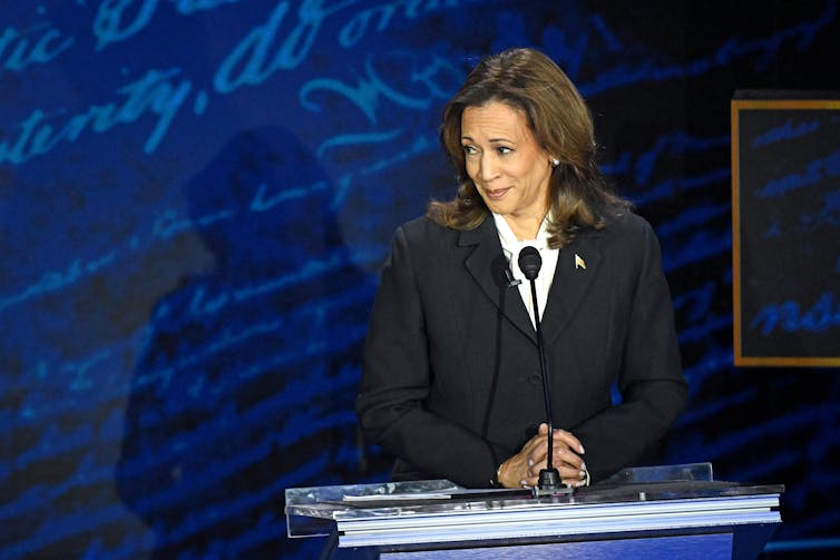 A woman wearing a black blazer stands at a podium and makes a face that suggests disbelief or amusement.