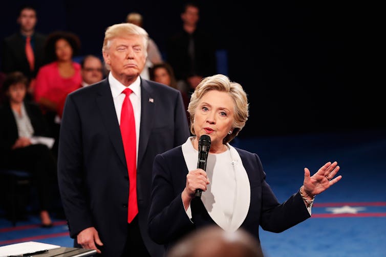 A blonde woman in a white shirt and black blazer speaks into a microphone, standing in front of a man with a dark suit and red tie looking toward her.
