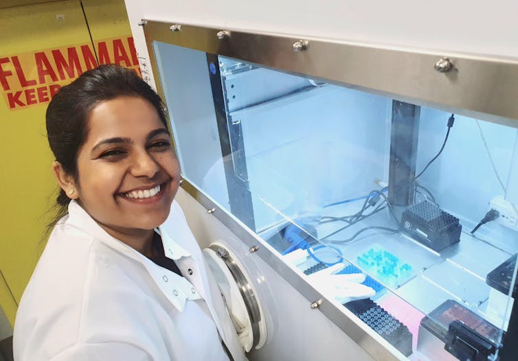A researcher in a white lab coat smiles at the camera next to a fume hood containing plates with small tubes.