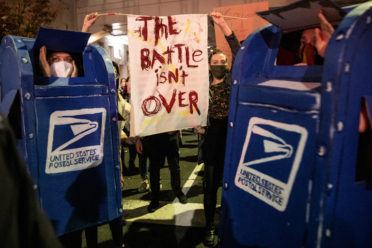 Demonstrators wear USPS mailbox costumes while people in background hold signs that read 'The battle isn't over'