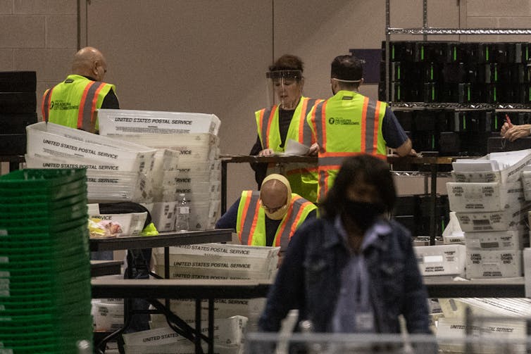Adults wearing flourescent yellow vests are surrounded by U.S. Postal Service plastic bins