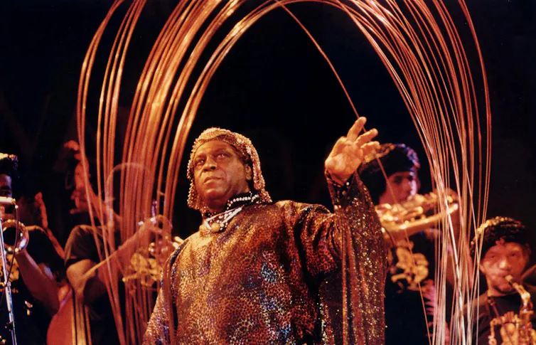Older man wearing elaborate sequined garb and headdress performs with musicians