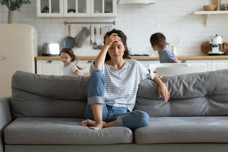 A tired woman sits on a couch kids run around behind her