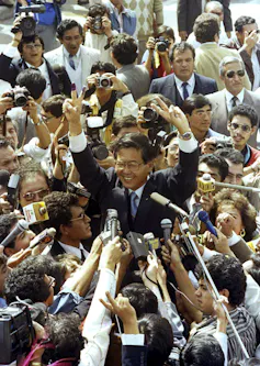 A man smiles and waves his hands as he is surrounded by people holding microphones.