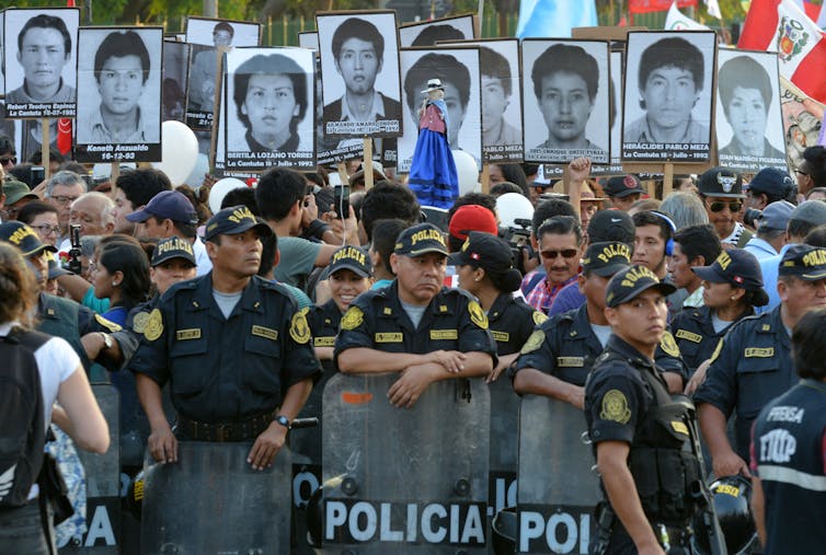 A group of people stand behind a lione of police. They hold aloft photos of people