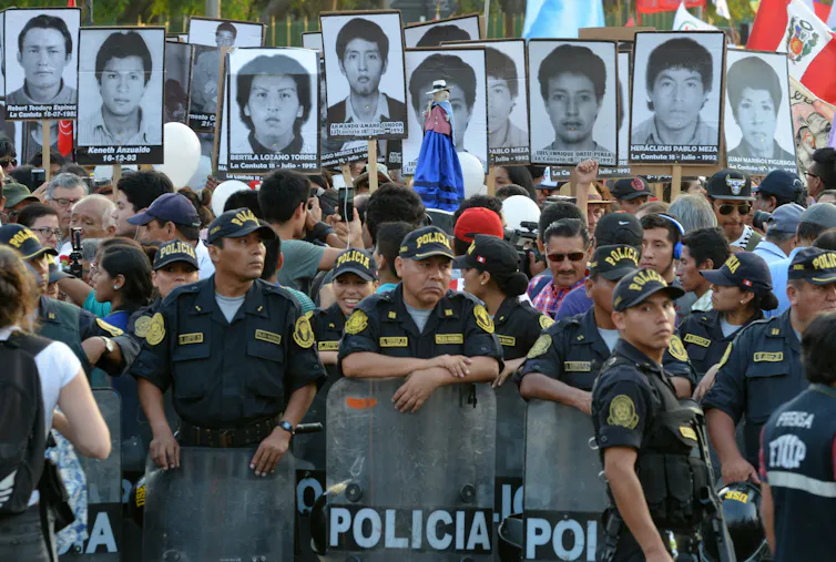 A group of people stand behind a lione of police. They hold aloft photos of people