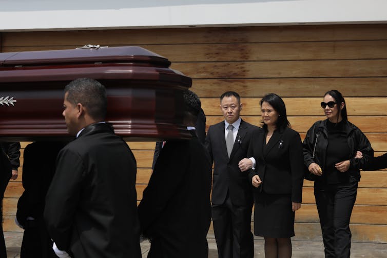 People dressed in black stand behind a coffin.