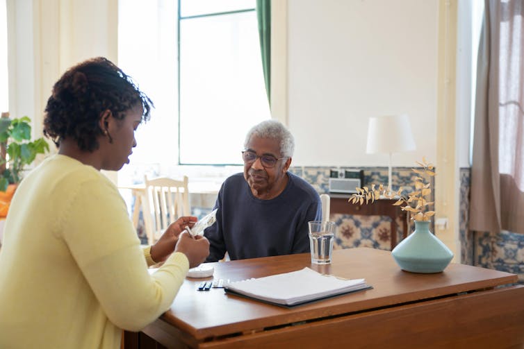A woman sitting at a table with a senior man, looking at a pill packet.
