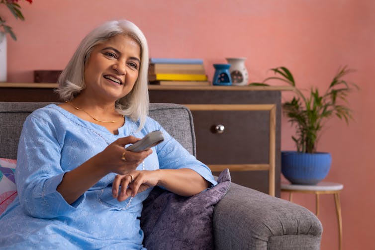 Uma mulher de meia-idade ou mais velha, com cabelos grisalhos, sentada no sofá em uma sala de estar colorida, sorrindo e apontando o controle remoto da televisão