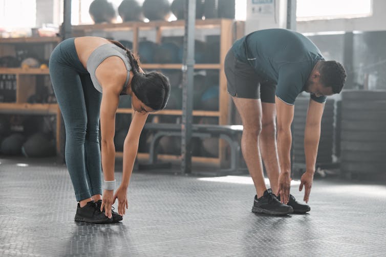 A young man and a young women stretch their legs in the gym by touching their toes.