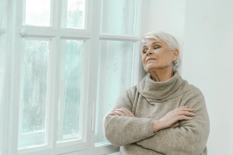 older woman in a thick jumper looking out of a window