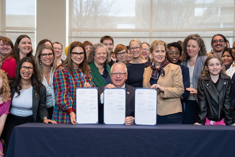 Walz sits smiling at a desk, surrounded by a crowd of smiling people