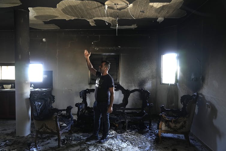 A man wearing black clothes reaches his hands up as he stands inside a charred room with two windows.