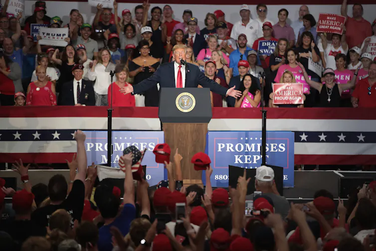 Trump stands at a podium in front of a large crowd and signs reading 'promises kept'