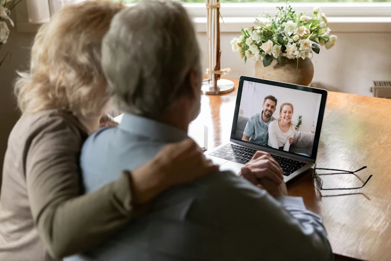 Two older people sit at a table video chatting with a younger couple on a laptop.
