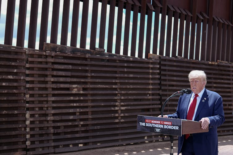 A middle-aged white man stands behind a podium near a tall wall.