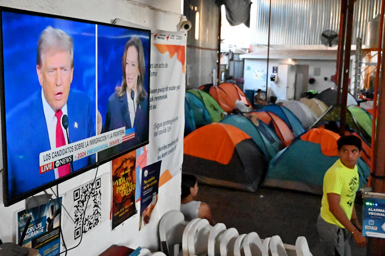 A young boy walks near rows of tents as he watches a television screen that has a white man wearing a business suit and a Black woman in a debate.