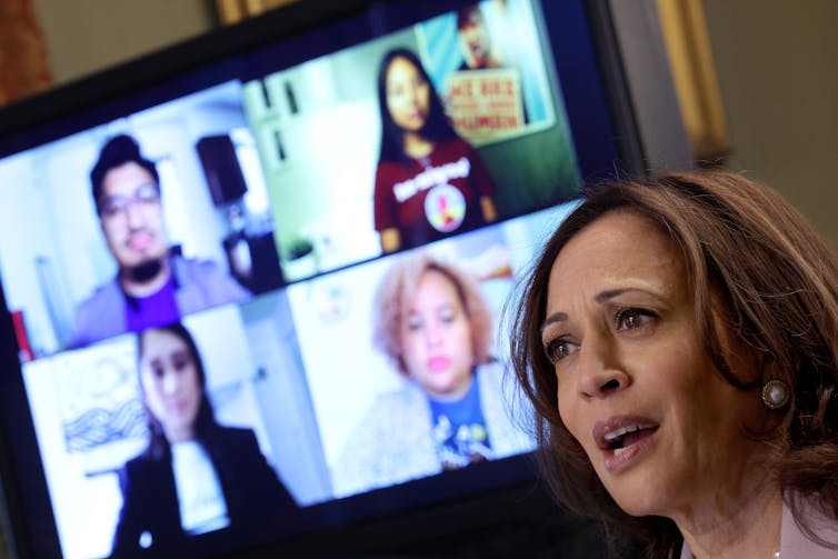 A Black woman is seen talking during a meeting where participants are on television screens in the background.