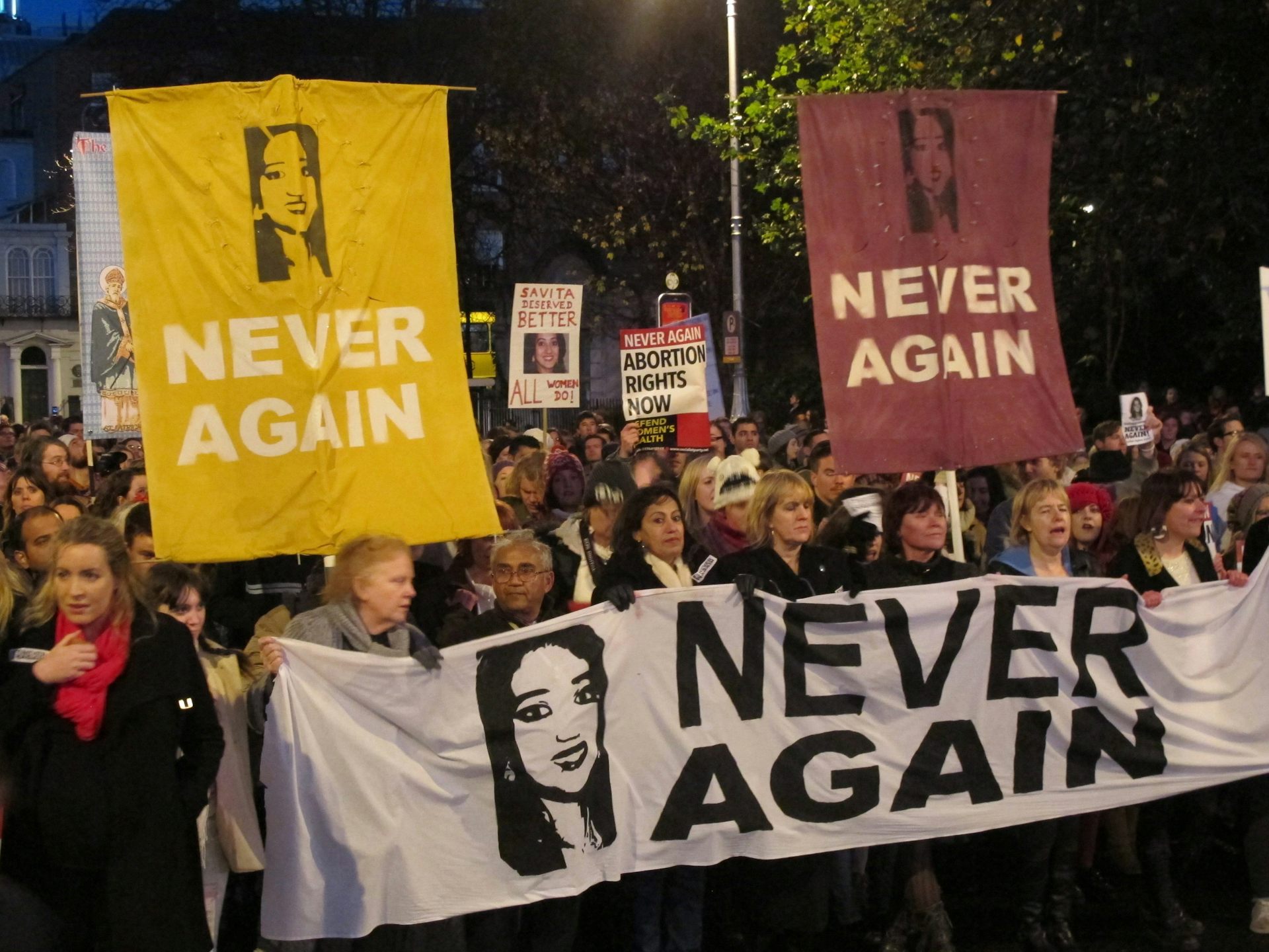 Protesters carry banners that read Never Again with a photo of a dark-haired woman's face.