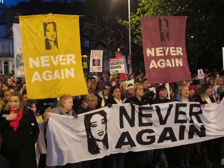 Protesters carry banners that read Never Again with a photo of a dark-haired woman's face.