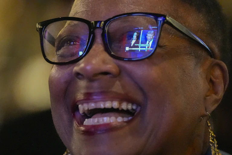 The televised debate between Donald Trump and Kamala Harris is reflected in a woman's glasses as she smiles while watching.