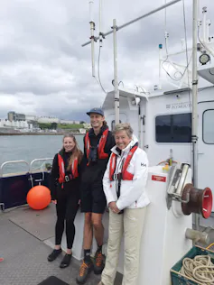 Three people wearing lifejackets on a boat at sea (woman, man, woman)