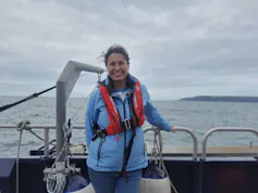 Woman in blue jacket and red lifejacket standing on boat at sea, grey sky