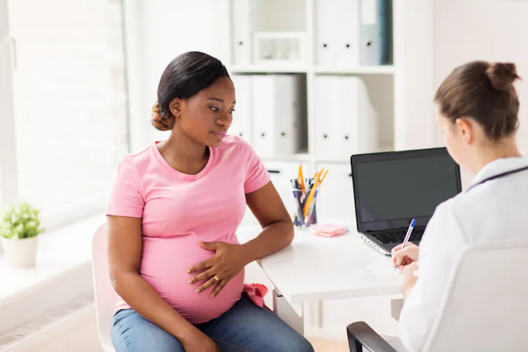 A pregnant woman talks to a doctor who writes on a notepad.
