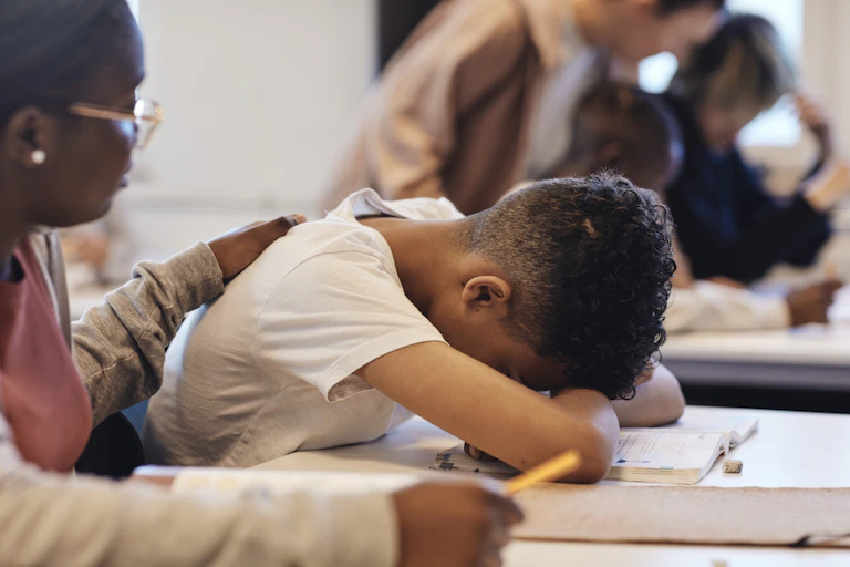 A boy puts his head down on his desk while a girl comforts him by placing her hand on his back.