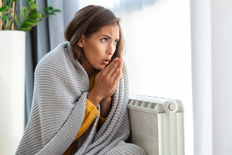 woman in grey blanket huddled over radiator, looks cold