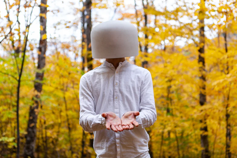 Un hombre vestido de blanco en un bosque extiende las manos con la cara tapada.
