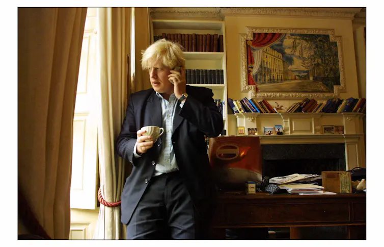 A younger Boris Johnson in front of a stocked bookshelf, desk and fireplace in his Spectator editor office, speaking on the phone and holding a cup of tea while looking out the window.