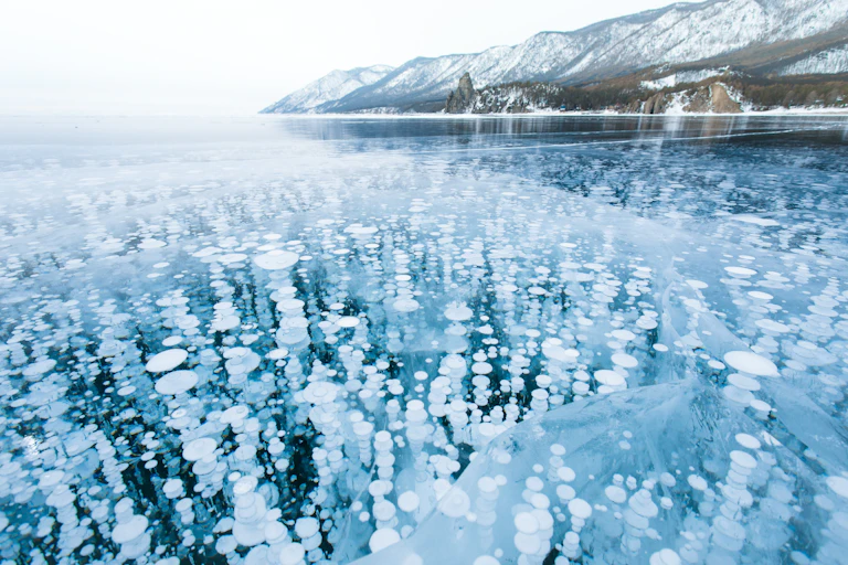 frozen lake with white bubbles, snowy mountains in background white sky