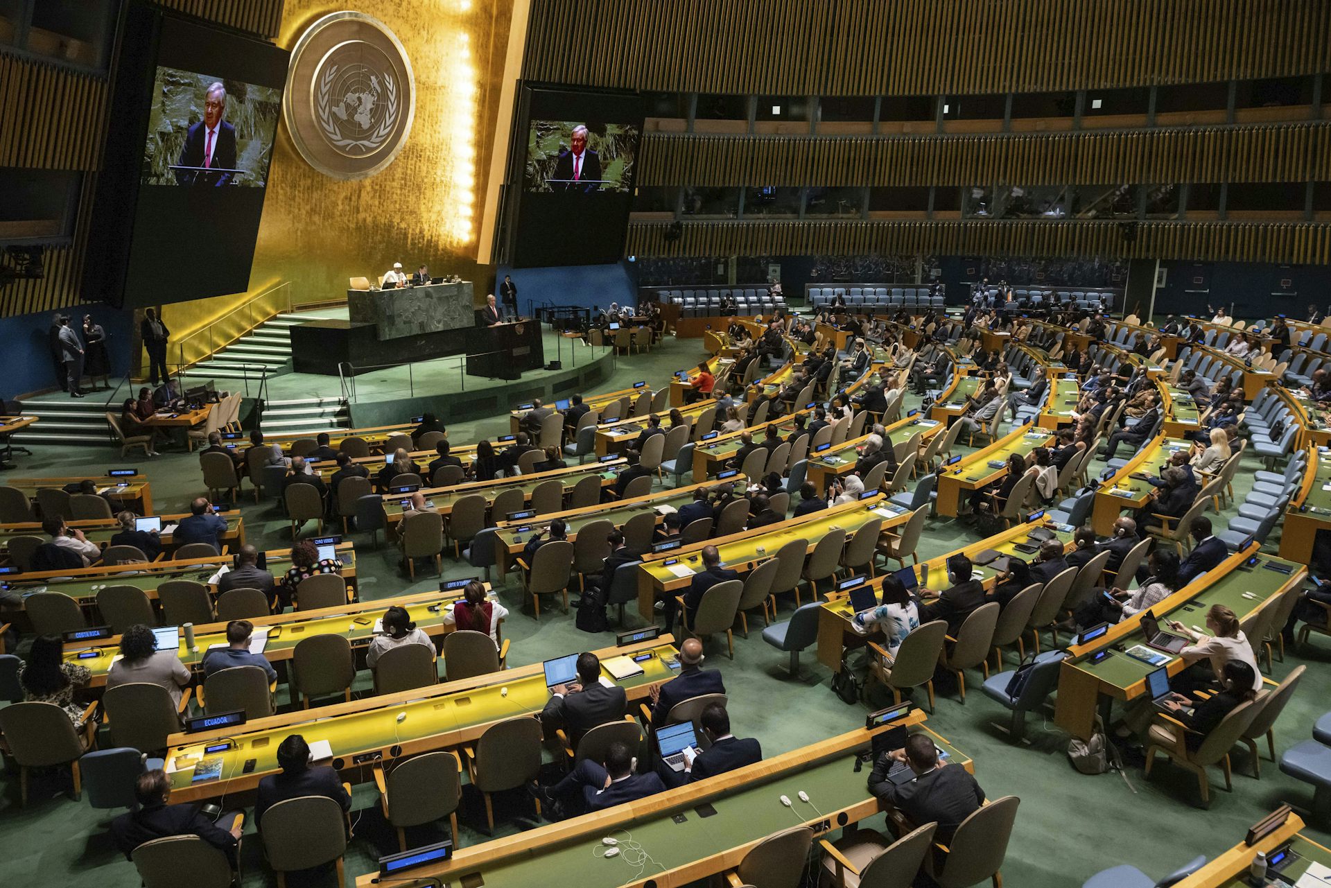 Un grand auditorium avec le logo de l'ONU sur un mur doré à l'avant.