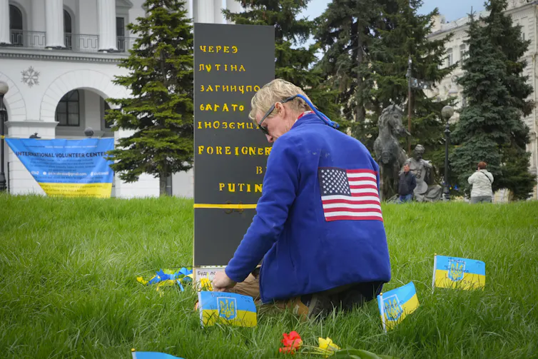 a blonde man wearing a blue jacket with the U.S. flag on the back kneels among grass surrounded by small Ukrainian flags