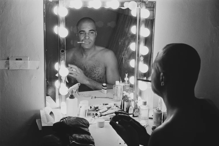 Shirtless young Black man smokes a cigar and winks while looking at his reflection in a mirror.