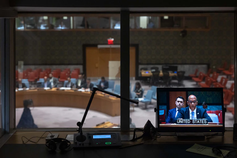 A small TV screen has a man talking with 'United States' written underneath. Through a window, seats are seen.
