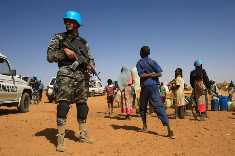 Man in camouflage uniform and blue helmet, holding a rifle, standing near a group of civilians including children