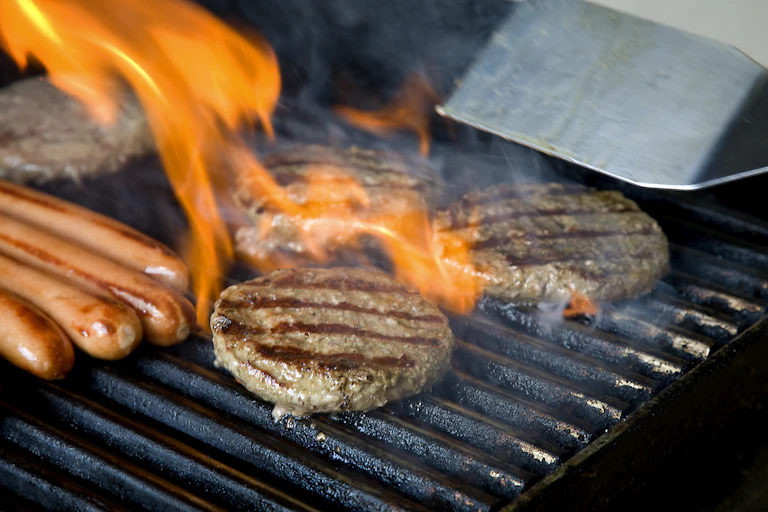 Beef patties and hot dogs on a hot grill with flames leaping around.
