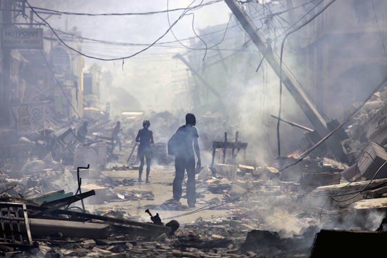 Two figures walk down a street in ruins amid smoke
