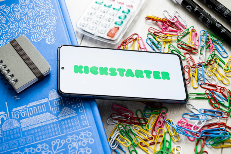 The Kickstarter logo is seen displayed on a smartphone on a desk covered with paper clips and other office supplies.
