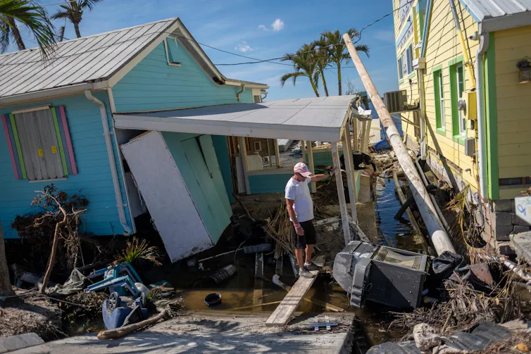 A man stands on porch of a house that is leaning precariously, with shreds of wood and a broken palm tree laying in the yard.
