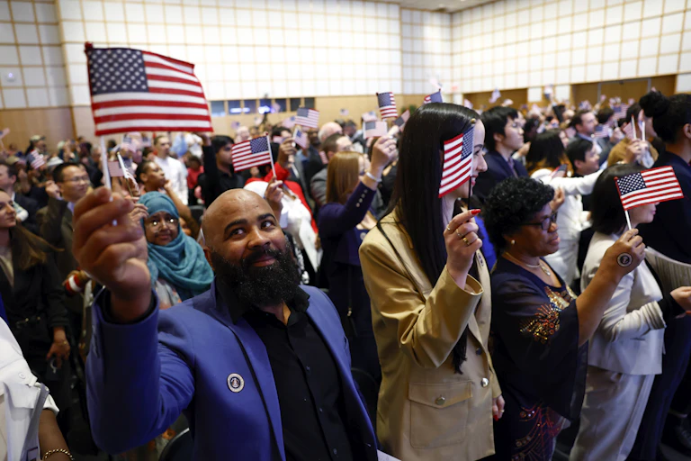 A large crowd of people waving small American flags are crowded into a small room. In the front left corner, a man wearing a purple jacket smiles.