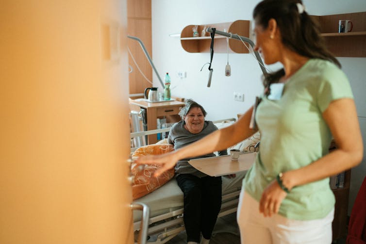 A nurse is looking at a smiling elderly woman sitting in bed in a hospital or aged care facility.