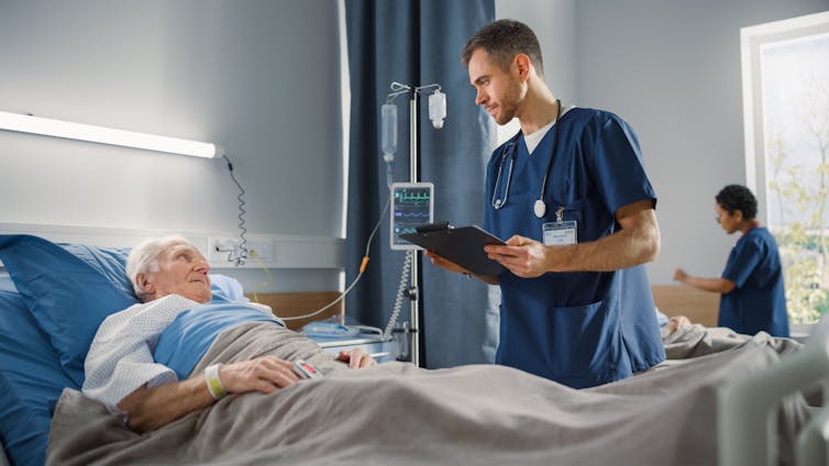 A male nurse talks to a senior man in a hospital bed.