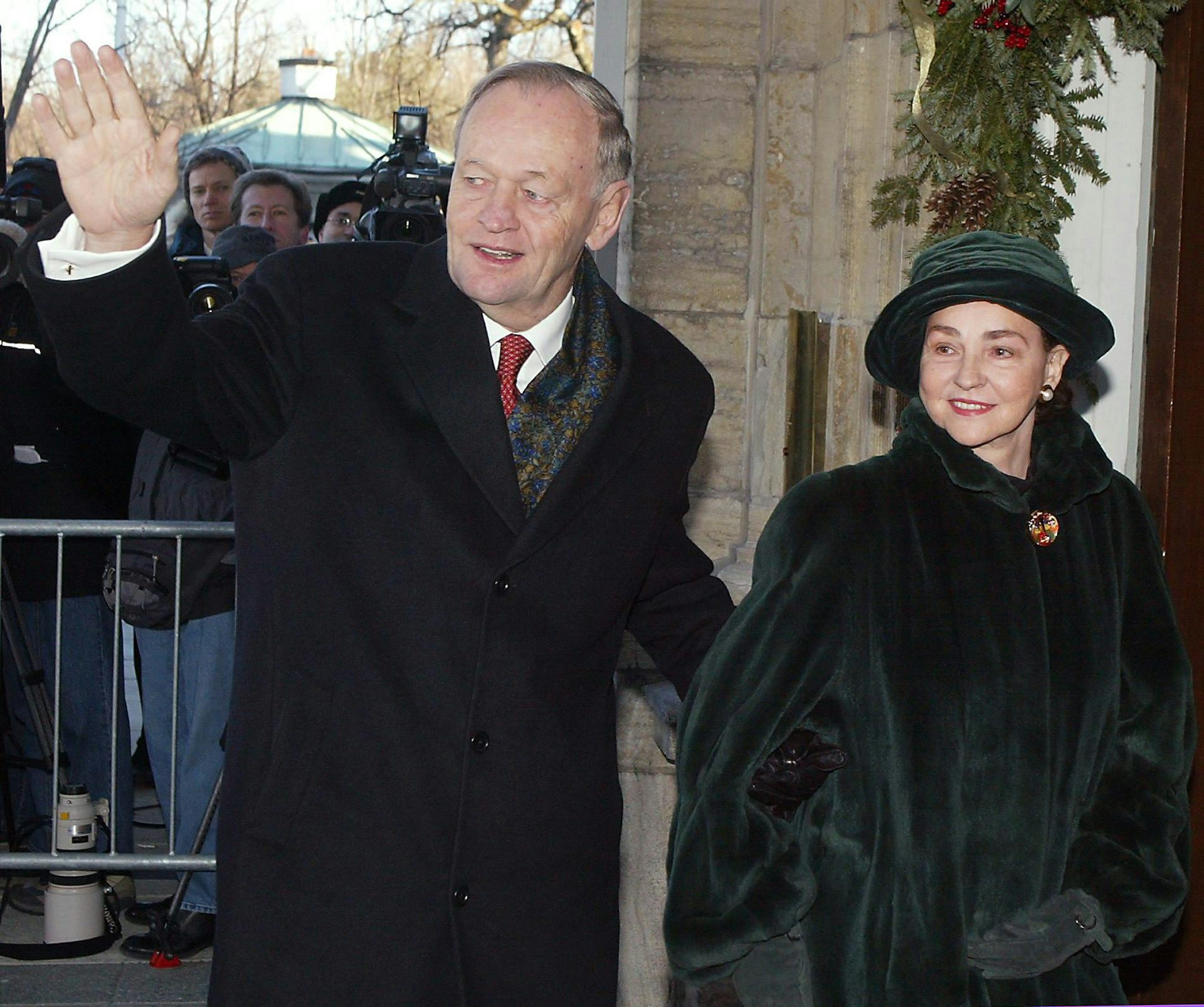 A man in a black wool coat waves next to a woman in a dark green coat and hat.