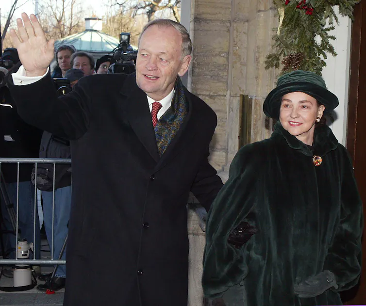 A man in a black wool coat waves next to a woman in a dark green coat and hat.