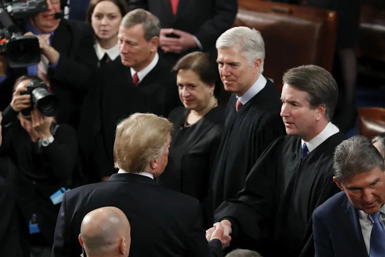 A line of simply dressed men and women wait to shake hands with another man who is facing away from the camera.