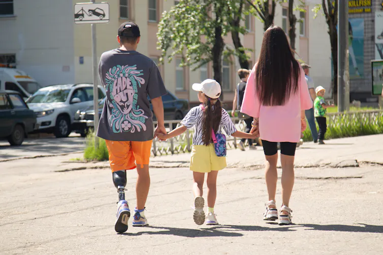 A casually dressed man with a prosthetic leg walking with a woman and child, seen from behind.
